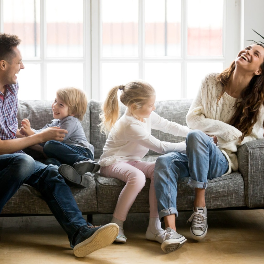 Happy parents and kids having fun tickling sitting together on sofa, cheerful couple laughing playing game with little active children son and daughter in living room at home, family funny activity