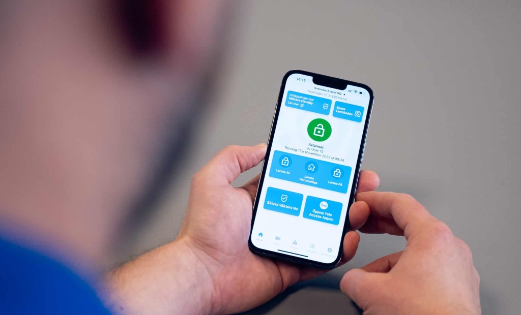 En person håller en mobiltelefon som visar vår säkerhetsapp. Personen syns svagt i bakgrunden. Appen på skärmen visar hur du enkelt kan hantera alla dina larmenheter och integrera med tjänster som Yale. Bilden visar även hur man kan hantera ett larm i sin app.